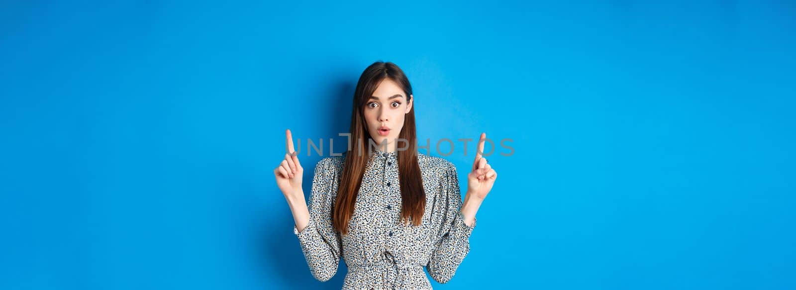 Excited pretty lady in dress say wow, looking intrigued by advertisement, pointing fingers up at logo, standing against blue background.