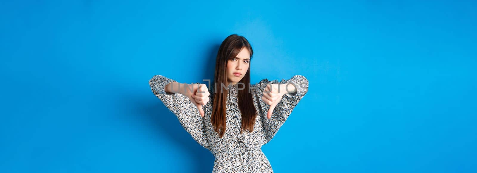 Negative emotion. Disappointed frowning woman with long hair and dress, showing thumbs down and grimacing with dislike, express aversion, standing on blue background.