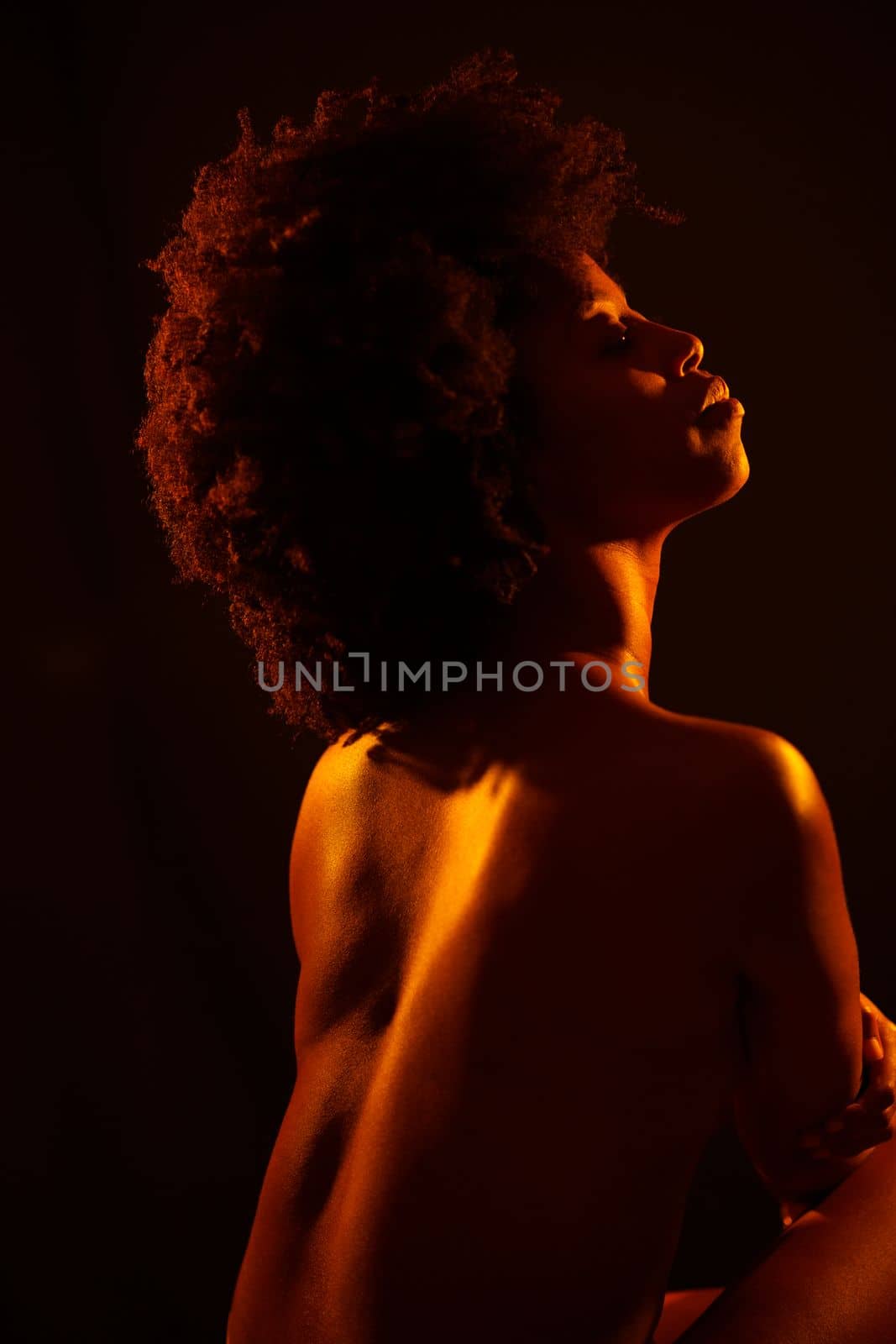 Sensual topless African American female model embracing body and closing eyes under orange neon illumination against black background.