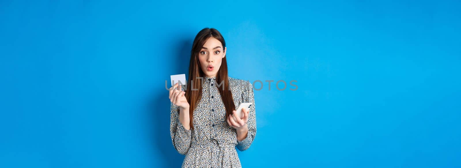 Online shopping. Excited girl paying with credit card on smartphone app, look amused, standing on blue background by Benzoix