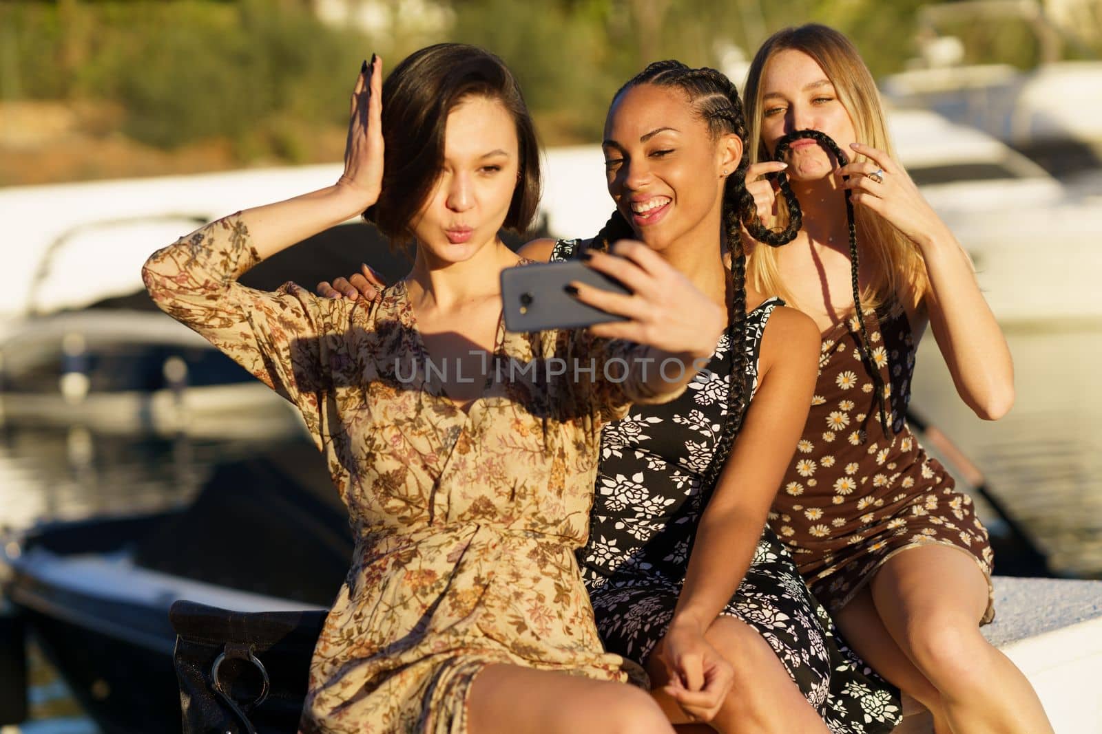 Delighted diverse female friends in dresses taking self portrait on smartphone while sitting together in wharf near river with boats