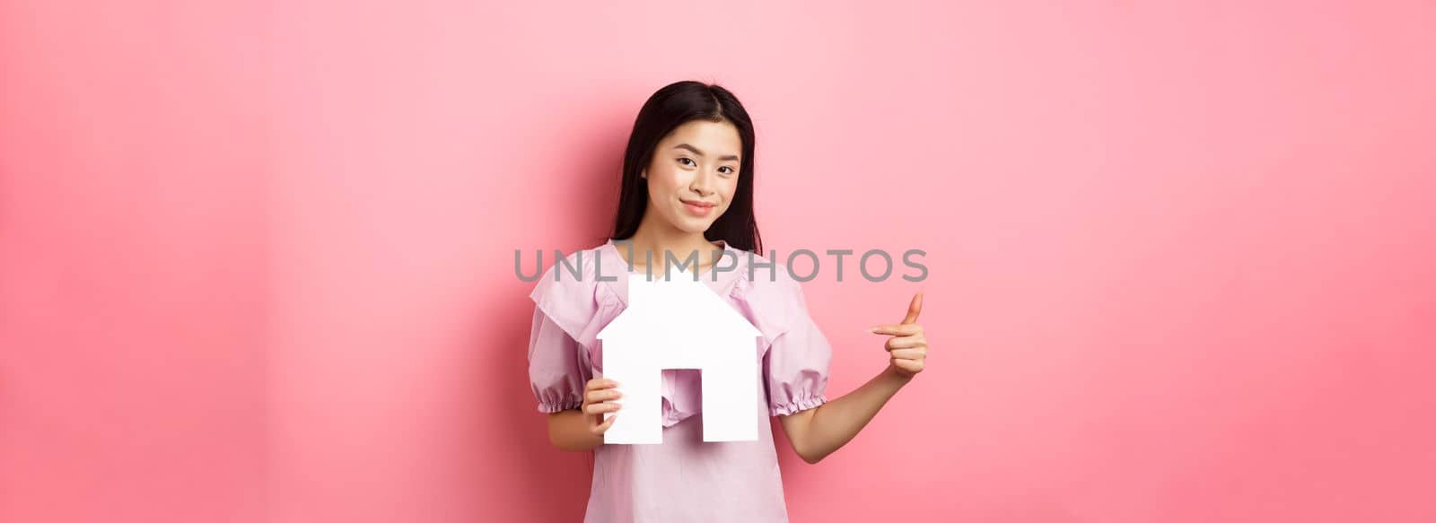 Real estate and insurance concept. Beautiful asian woman pointing at paper house cutout, showing agency logo, standing in dress on pink background.