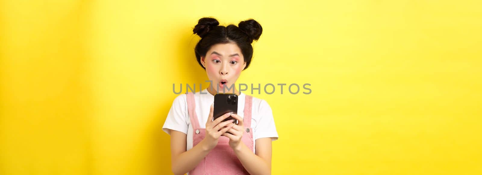 Asian teen girl look excited at smartphone screen, reading news on phone, standing on yellow background.