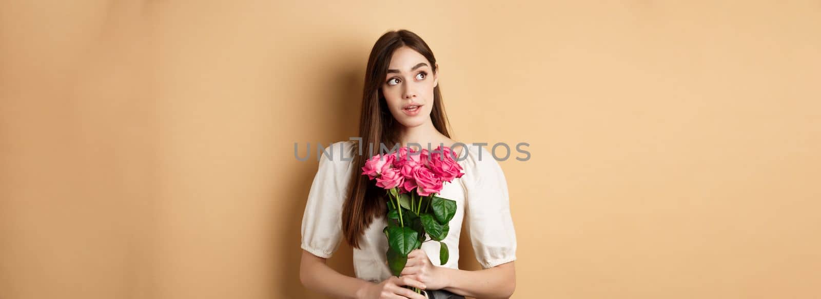 Romantic Valentines day concept. Dreamy woman with bouquet of pink rose, looking aside at logo, holding flowers on beige background by Benzoix