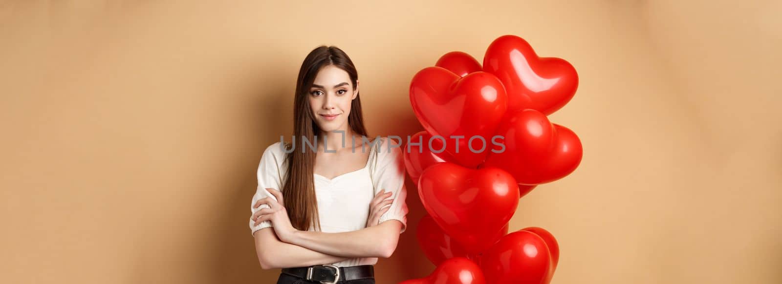 Sassy young girl cross arms on chest and smiling confident, standing near Valentines day hearts balloons and looking at camera, beige background.