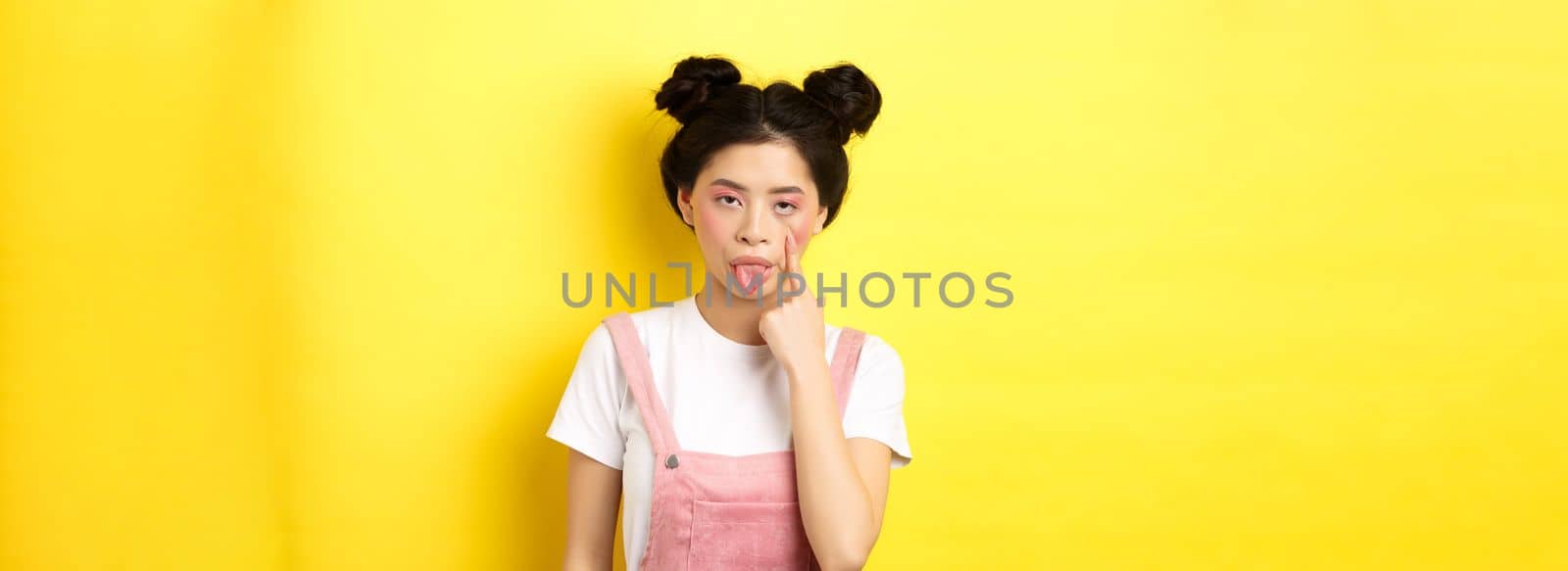 Rude asian girl stretch eyelid and showing tongue, mocking someone, standing on yellow background.