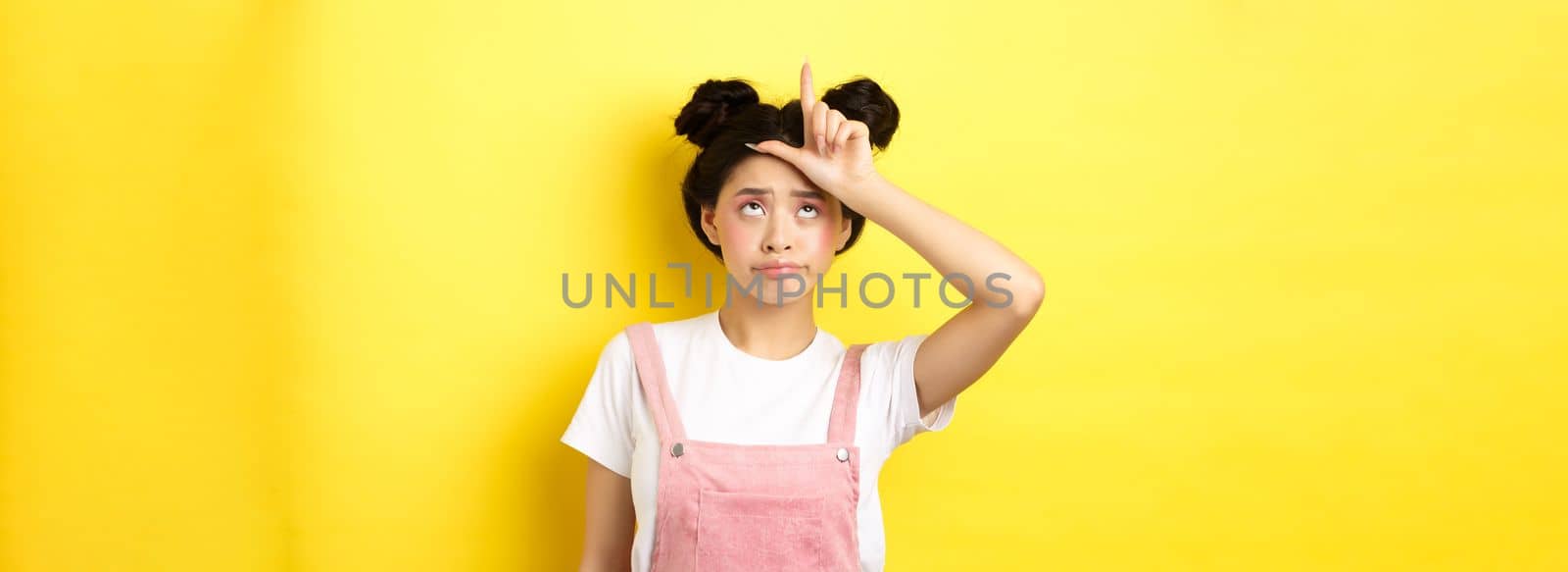 Sad girl showing loser sign on forehead and sulking upset, feeling disappointed in herself, standing on yellow background.