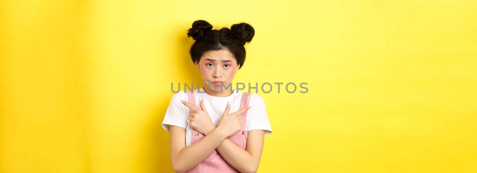 Indecisive timid asian girl pointing sideways, showing left and right ways and sulking upset, standing sad on yellow background.