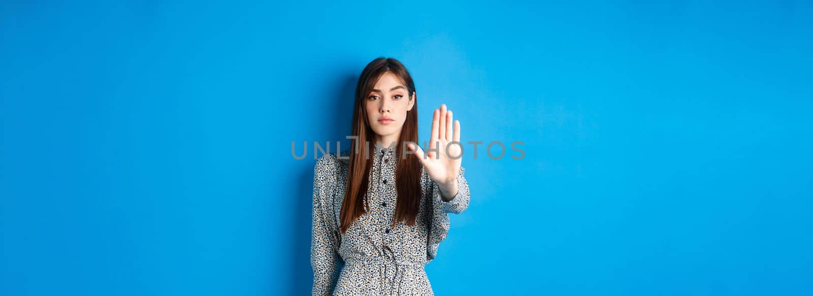 Serious and confident woman with long hair and dress, stretch out hand and say no, make stop gesture, prohibit bad action, standing on blue background.