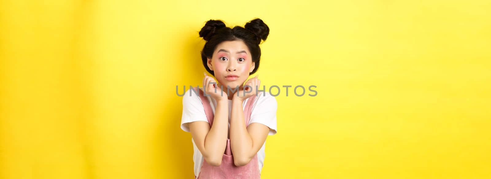 Cute teenage asian girl with beauty makeup, listen with interest, lean on hands and looking excited at camera, standing on yellow background.