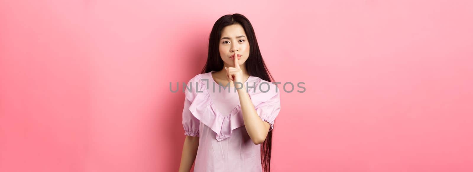 Cute asian girl tell to be quiet, scolding loud person, showing shush sign with finger pressed to lips, standing in dress on pink background by Benzoix