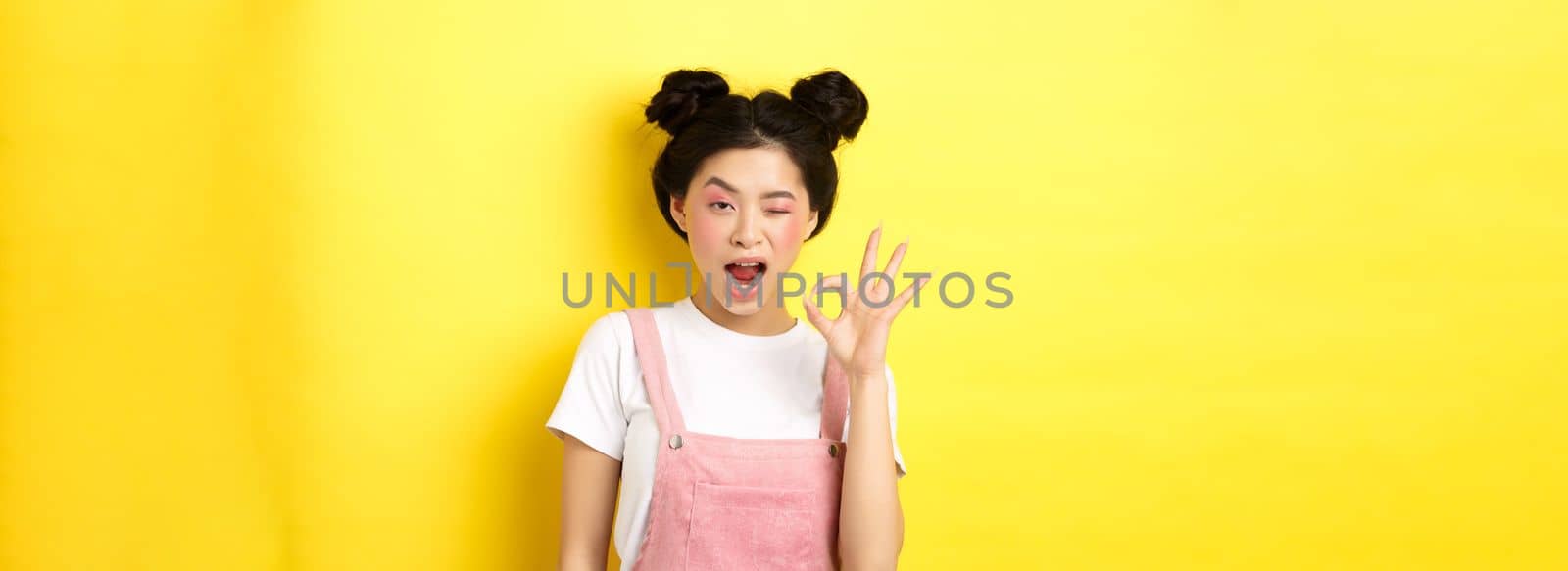 Alright. Confident asian female model with bright makeup and summer clothes, showing okay sign, winking and say yes, agree and approve, yellow background.