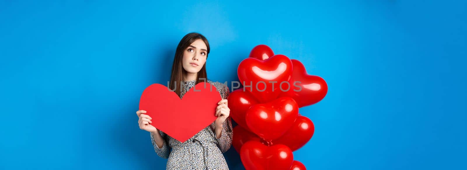 Valentines day. Dreamy pretty lady in dress, holding big red heart cutout and searching for true love, looking up pensive, standing near holiday balloons on blue background.