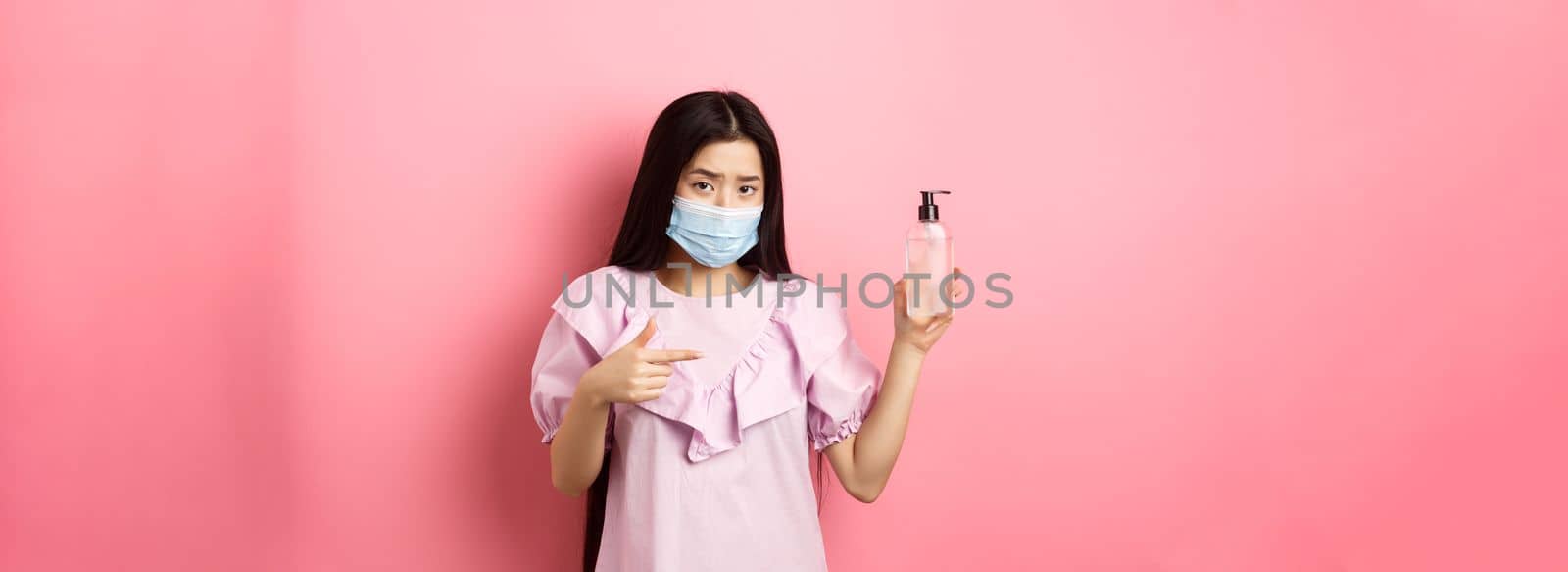 Healthy people and covid-19 pandemic concept. Skeptical asian woman frowning, wearing face mask, pointing at bottle of hand sanitizer, standing against pink background.