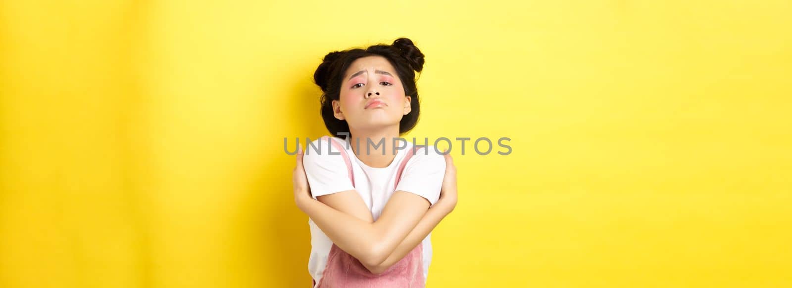 Lonely sad teen girl hugging herself, wanting to relationship and cuddles, standing alone on yellow background.