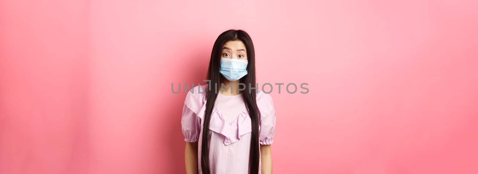 Covid-19, pandemic lifestyle concept. Surprised asian woman in medical mask look in awe, raising eyebrows with disbelief, standing against pink background by Benzoix