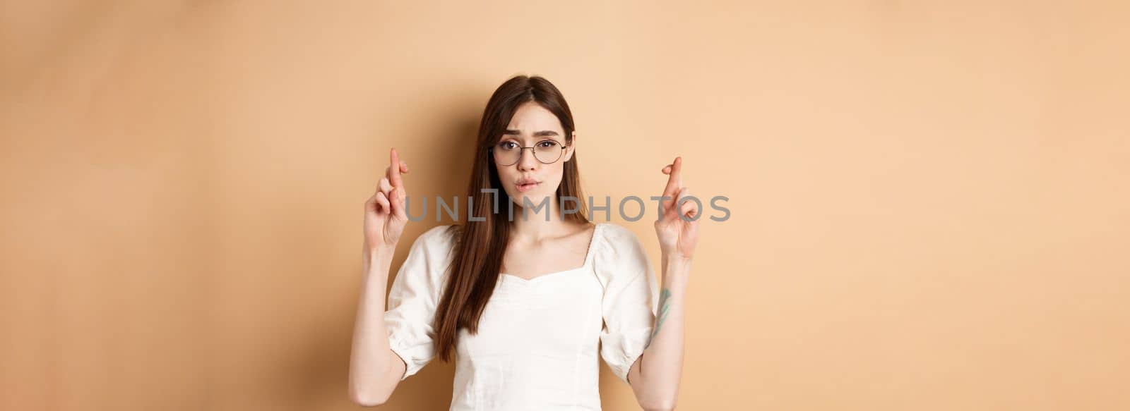 Hopeful girl in glasses cross fingers for good luck, biting lip nervous and looking at camera, waiting for results, praying or making wish, beige background.