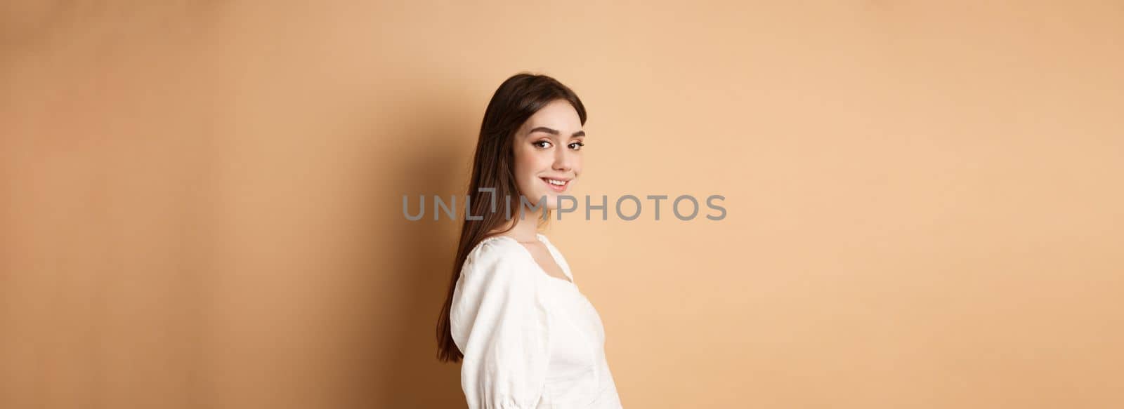 Profile of pretty european girl turn head at camera and smiling coquettish, standing in white blouse against beige background.