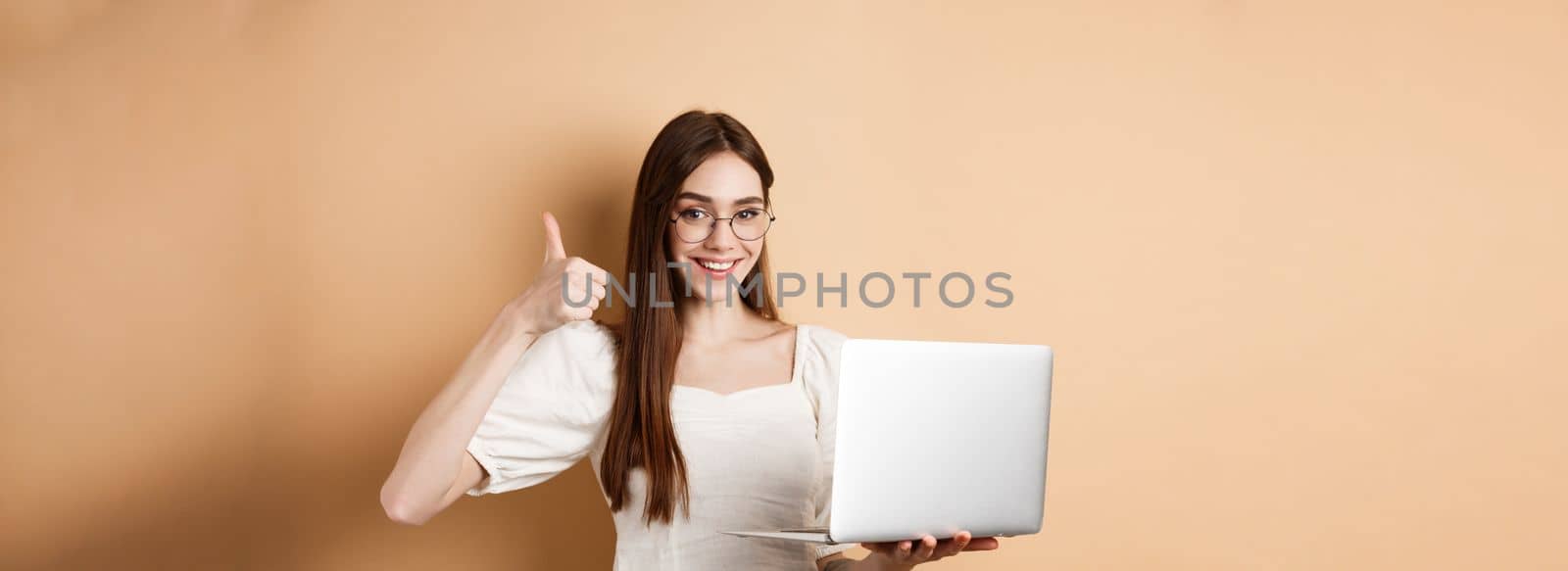 Smiling woman in glasses using laptop and showing thumb up, working on computer, standing on beige background.