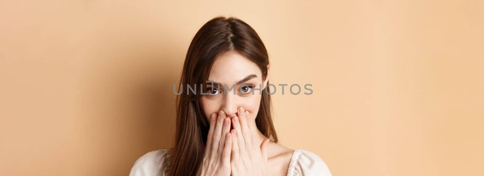 Close up of coquettish pretty girl covering mouth, looking happy and romantic, standing on beige background.