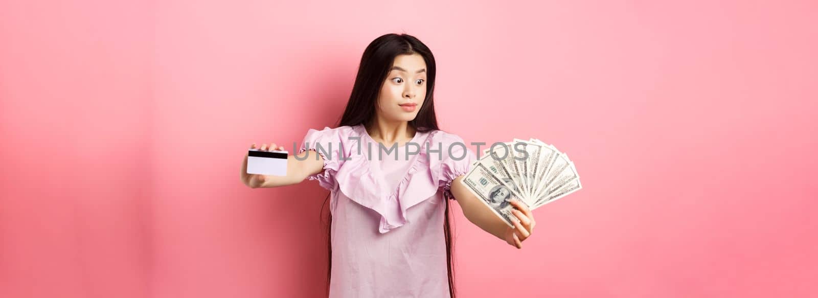 Excited teen asian girl stretch out hands with dollar bills and plastic credit card, look at money with amazement, standing on pink background.