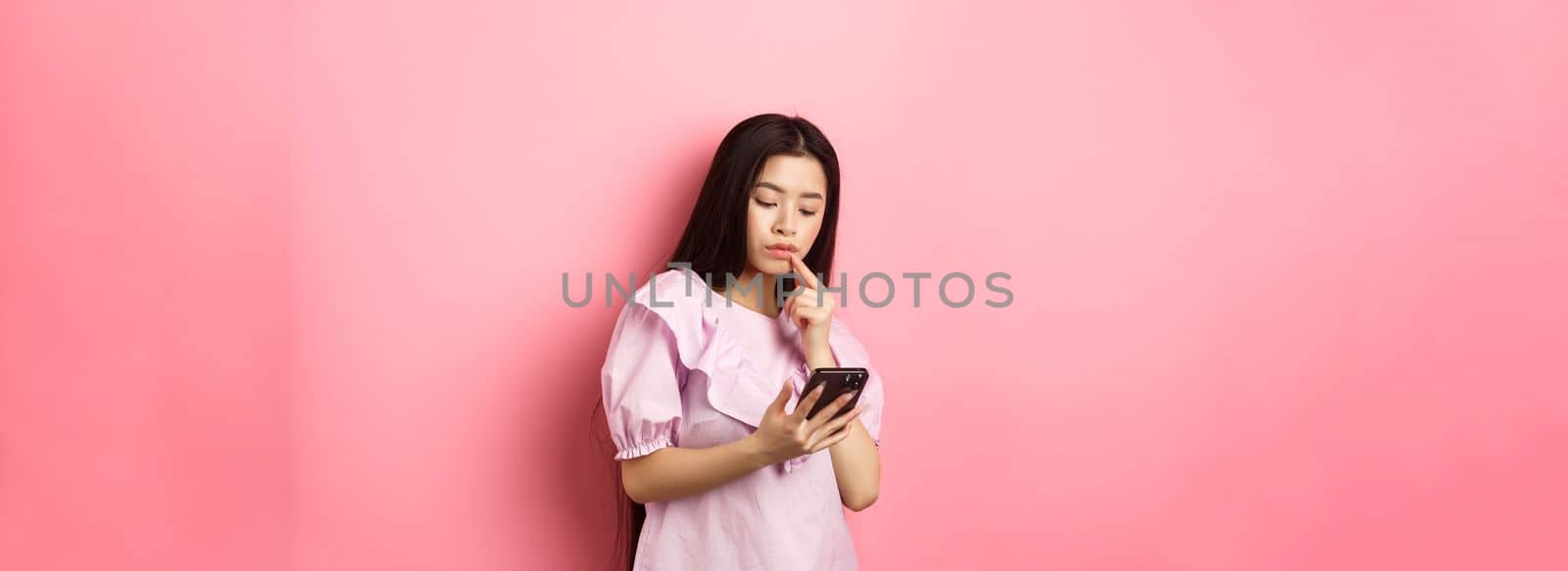 Online shopping. Pensive asian girl looking at smartphone screen, making choice, standing in dress against pink background.