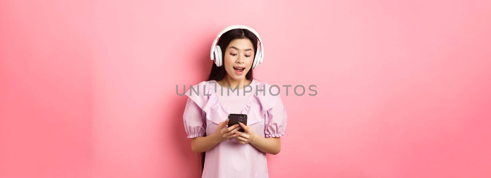Happy beautiful woman in wireless headphones watching video on smartphone, looking at phone amused, standing against pink background.