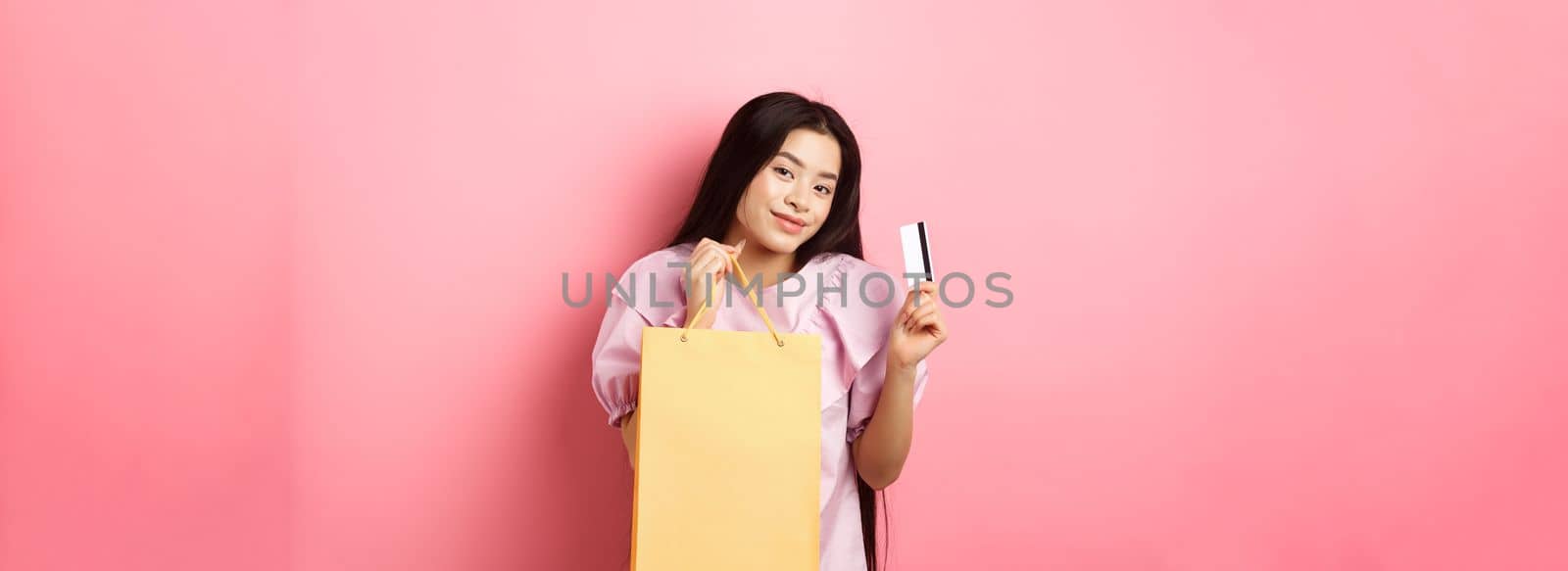Shopping. Stylish asian girl showing shop bag and plastic credit card, standing on pink background.