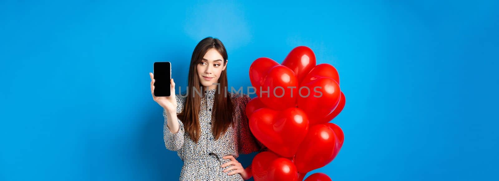 Valentines day. Pretty smiling woman in dress showing empty smartphone screen, standing near romantic balloons, blue background.