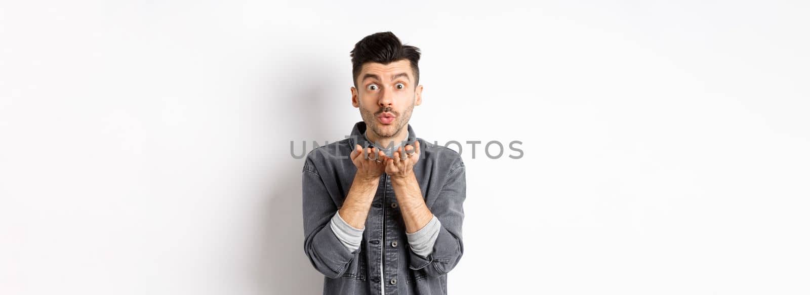 Romantic handsome man blowing air kiss, looking silly at camera, standing in trendy clothes on white background.