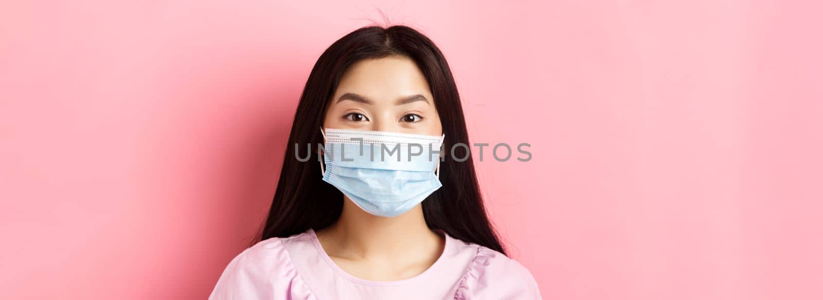 Covid-19 and healthy people concept. Close-up of asian cute girl wearing medical mask during virus outbreak, standing against pink background.