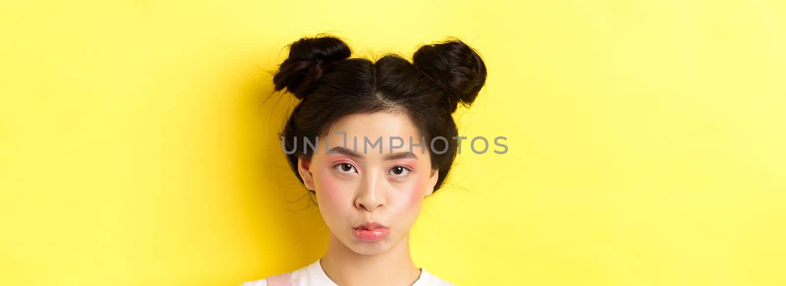 Close up portrait of moody asian girl pouting and looking upset at camera, standing wiht glamour makeup and hairbuns on yellow background.