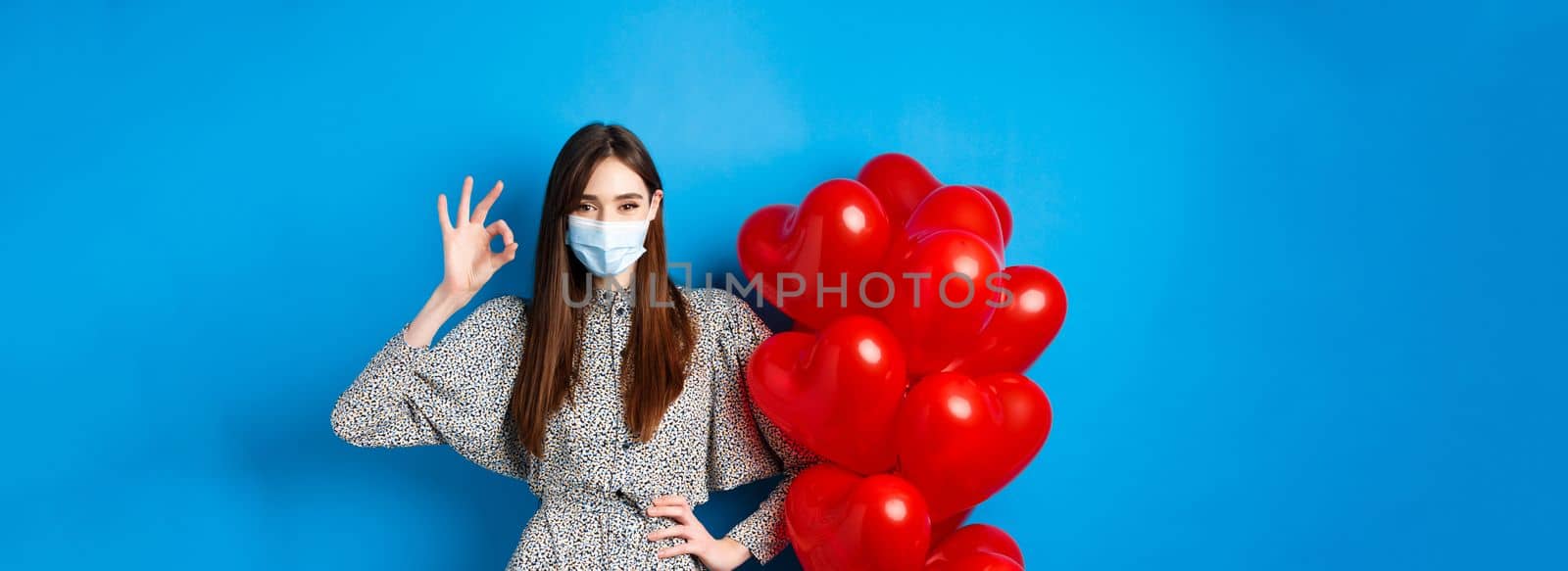 Covid-19, Valentines day and pandemic concept. Beautiful young woman in face mask and dress, showing OK sign in approval, standing near romantic red hearts balloons, blue background by Benzoix