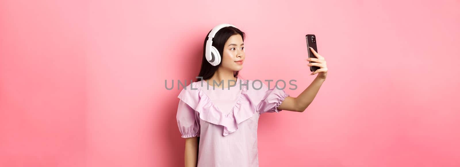 Beautiful japanese girl taking selfie on smartphone, wearing wireless headphones, standing against pink background.