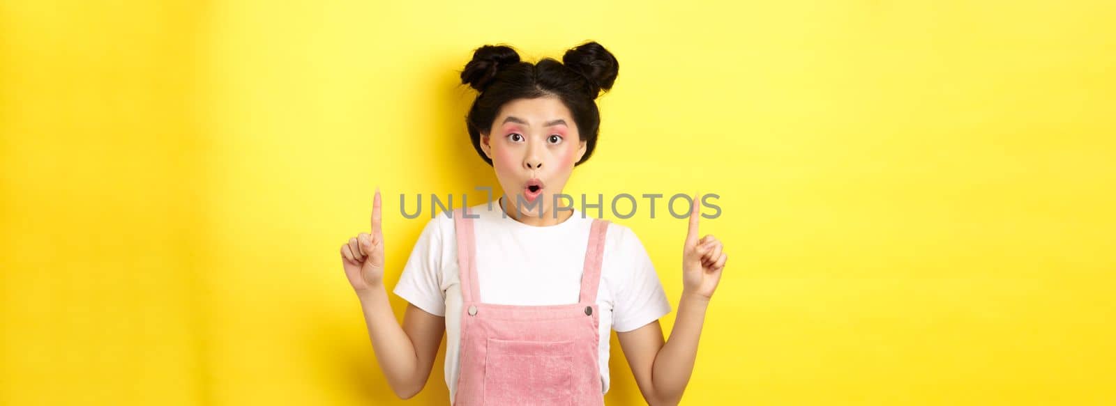 Excited asian female model with glamour makeup, pointing fingers up and say wow amazed, checking out promo deal, standing on yellow background by Benzoix