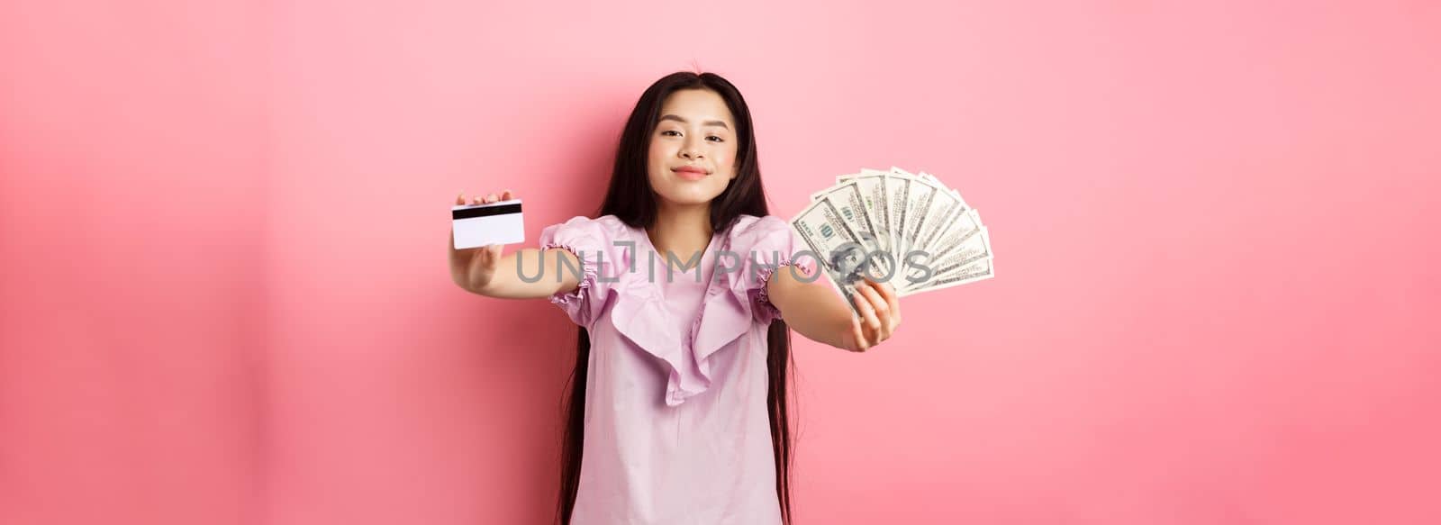 Beautiful asian woman showing plastic credit card and dollar bills, smiling pleased, standing against pink background by Benzoix