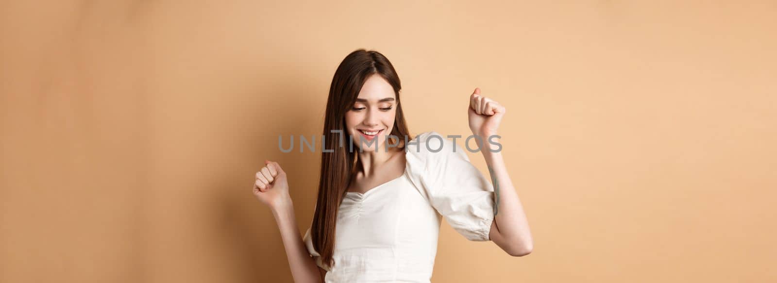 Carefree woman dancing and having fun, close eyes and smiling, standing on beige background.