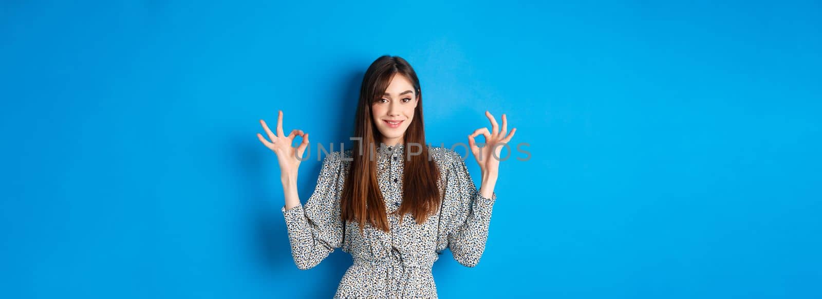 Confident adult woman in dress with natural hair color, showing okay signs and smiling, like and approve something good, standing on blue background by Benzoix