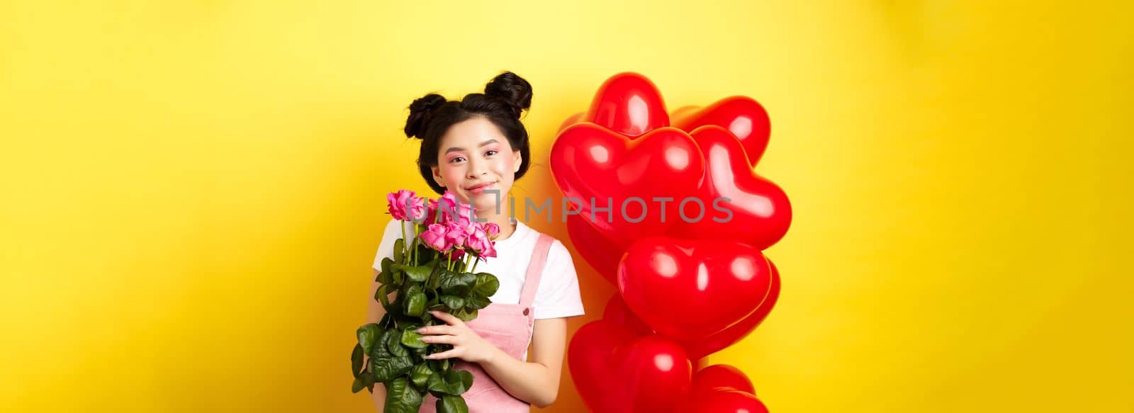 Happy Valentines day. Beautiful asian woman dressed for romantic date, holding bouquet of flowers and smiling. Girl with roses standing near heart balloons, yellow background by Benzoix