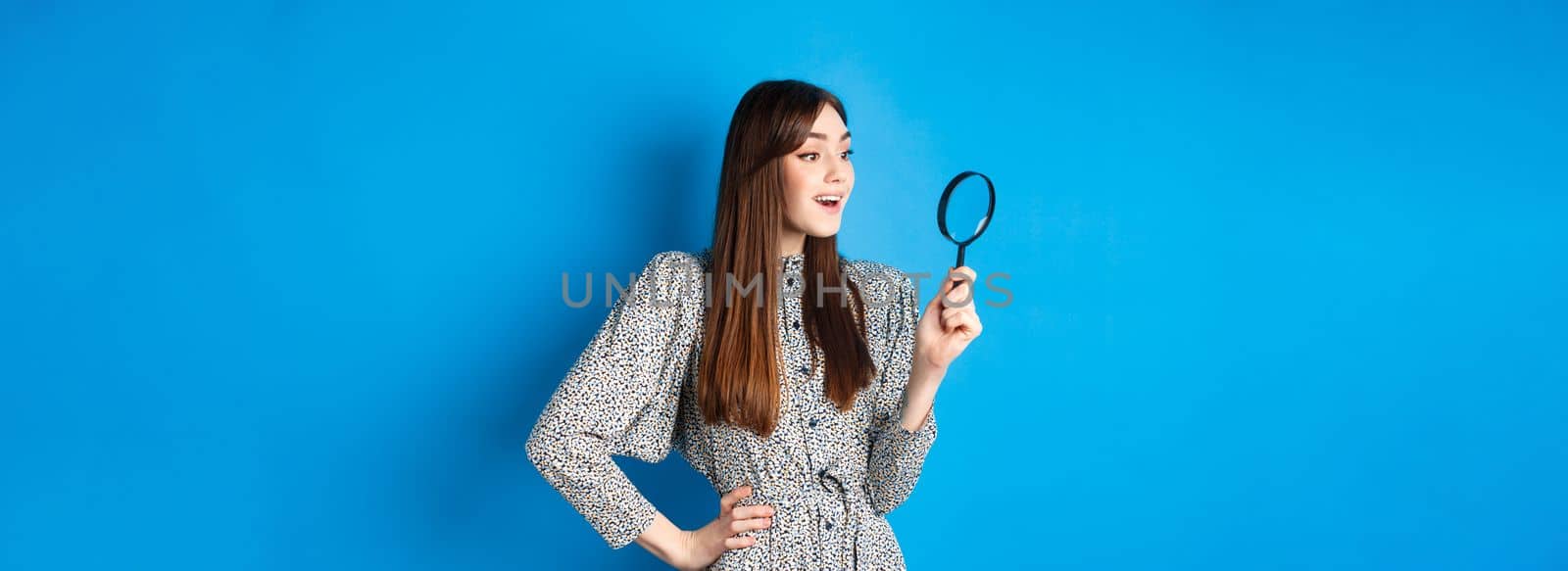 Excited girl looking left with magnifying glass, found interesting promo, investigating or searching, standing on blue background.