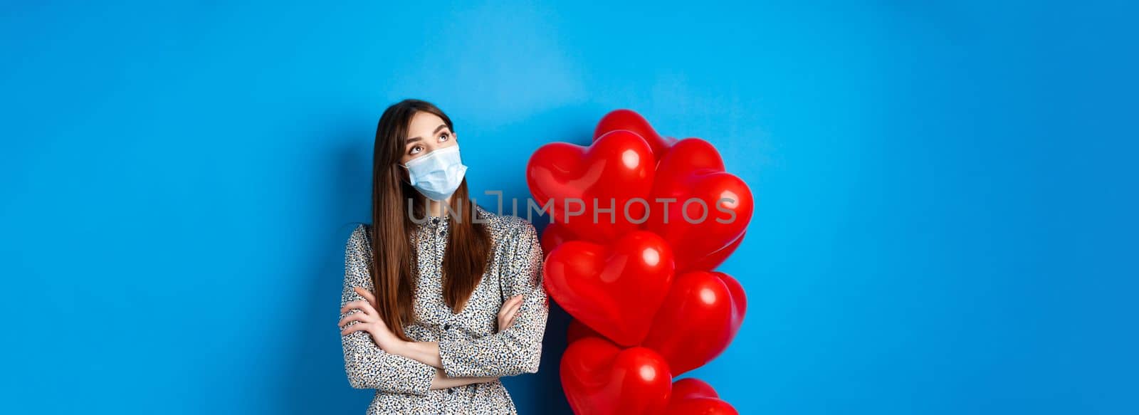 Covid-19, quarantine and health conept. Dreamy beautiful girl in face mask and dress, looking at upper left corner pensive, standing near Valentines day balloons, blue background by Benzoix