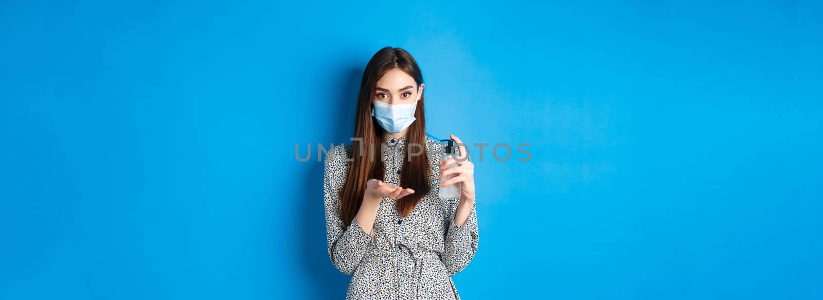 Covid-19, social distancing and healthcare concept. Beautiful girl in medical mask using hand saniziter, holding antiseptic bottle, standing on blue background.