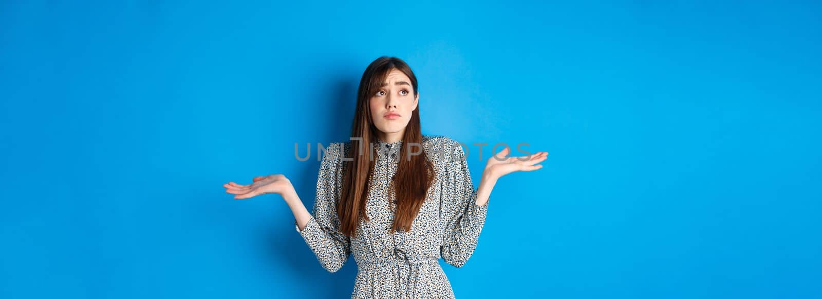 Confused pretty girl in dress shrugging shoulders and looking aside sad, know nothing, standing against blue background by Benzoix