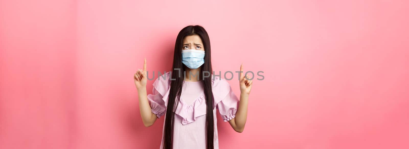 Coronavirus, quarantine and lifestyle concept. Disappointed sad asian girl pointing fingers up, frowning and sulking at unfair situation, standing against pink background by Benzoix