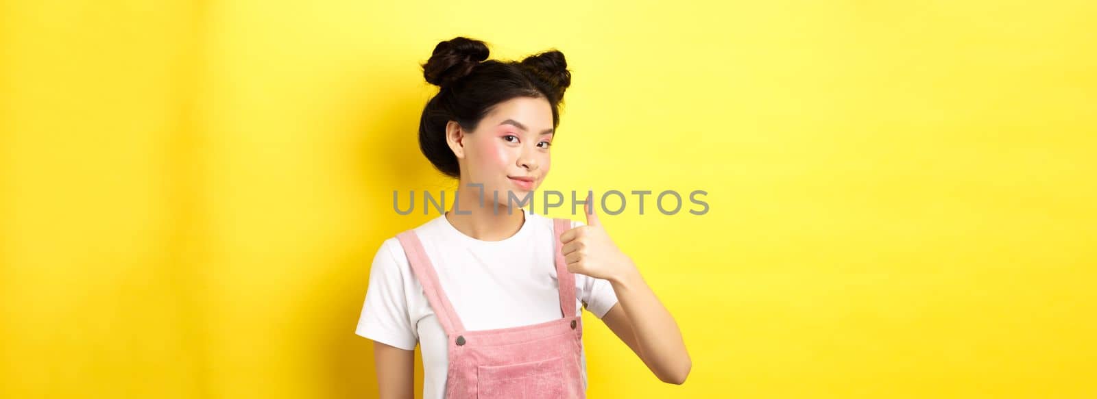 Beautiful asian girl with bright pink makeup, showing thumb up in approval and smiling satisfied, recommending promo, yellow background.