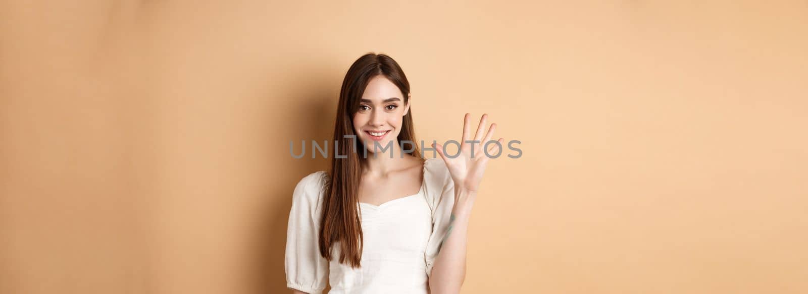 Attractive young woman show fingers number five, smiling and looking confident, standing on beige background.