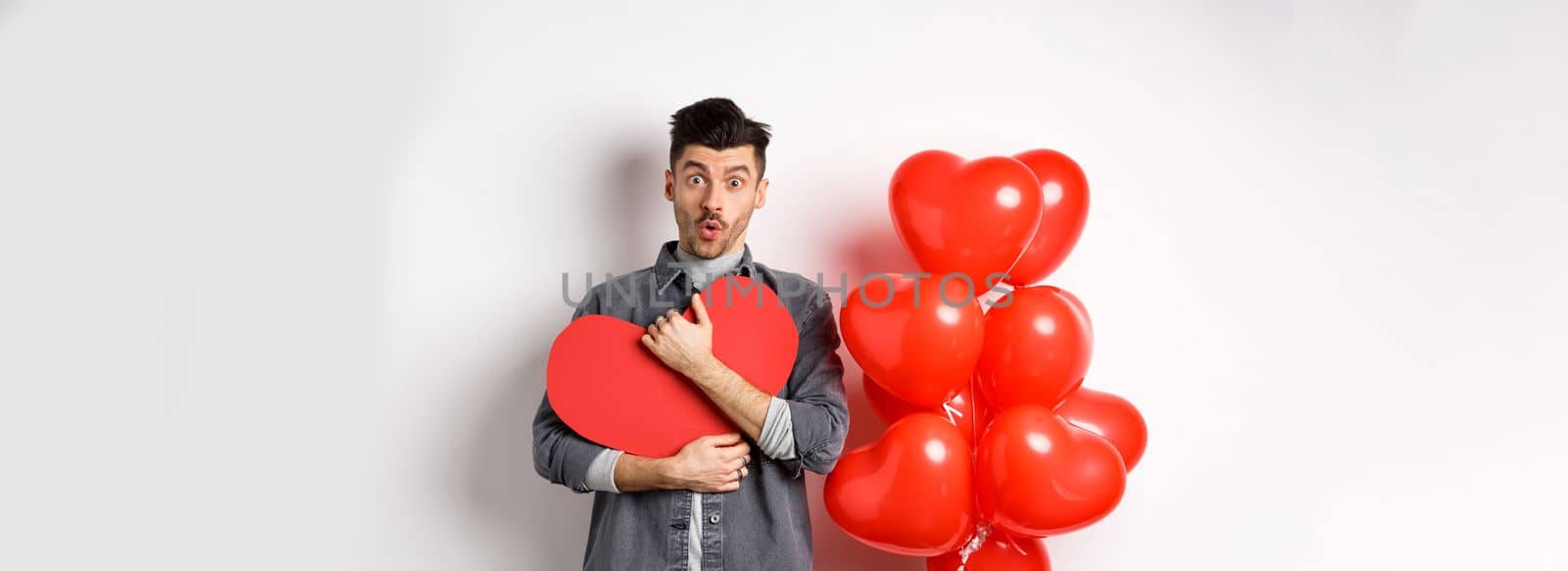 Surprised man holding Valentines heart card and saying wow, looking amazed at camera, receive secret confession on lovers day, standing near romantic balloons on white background.
