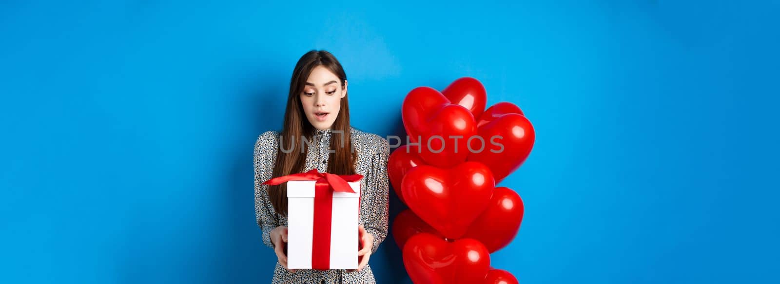 Valentines day. Image of beautiful girl looking surprised at gift box, receive present from lover, standing in dress on blue background by Benzoix