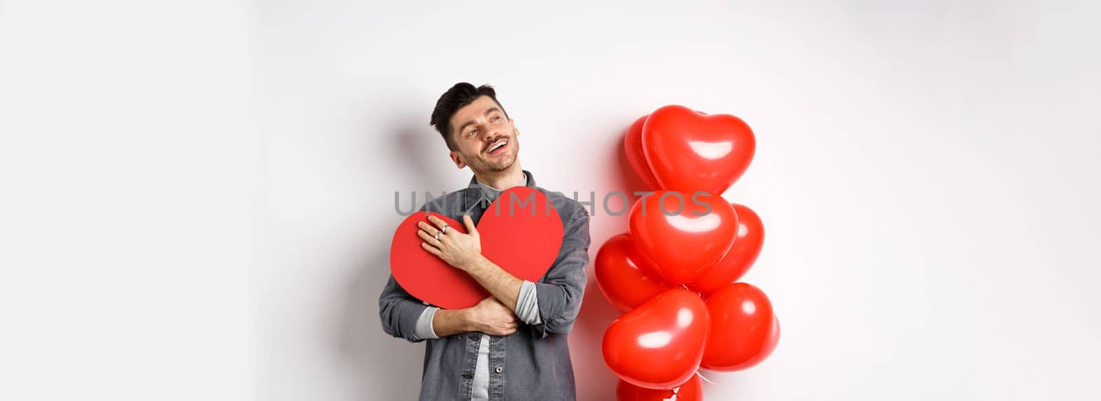 Romantic man hugging big red heart cutout and looking away dreamy, thinking of girlfriend and valentines day, imaging lover, standing on white background.