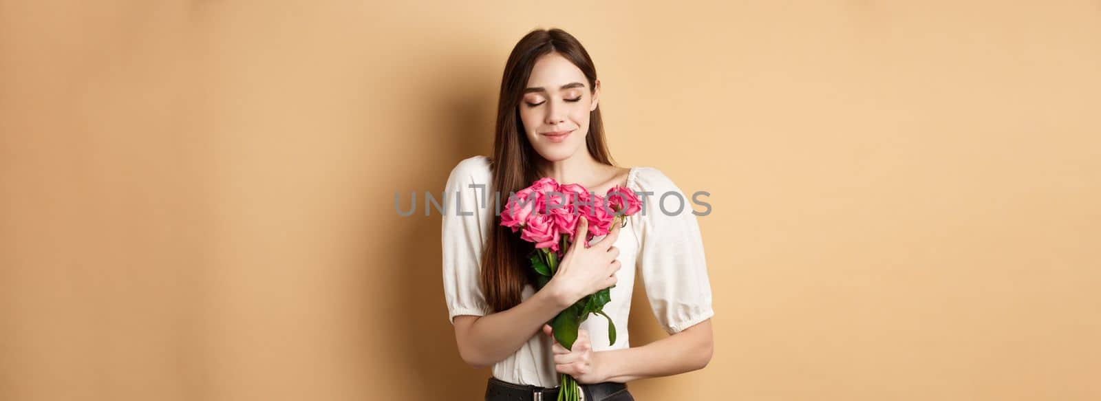 Valentines day. Tender and romantic girl, smell roses and smile with closed eyes. Girlfriend hugging gift flowers from lover, standing on beige background.